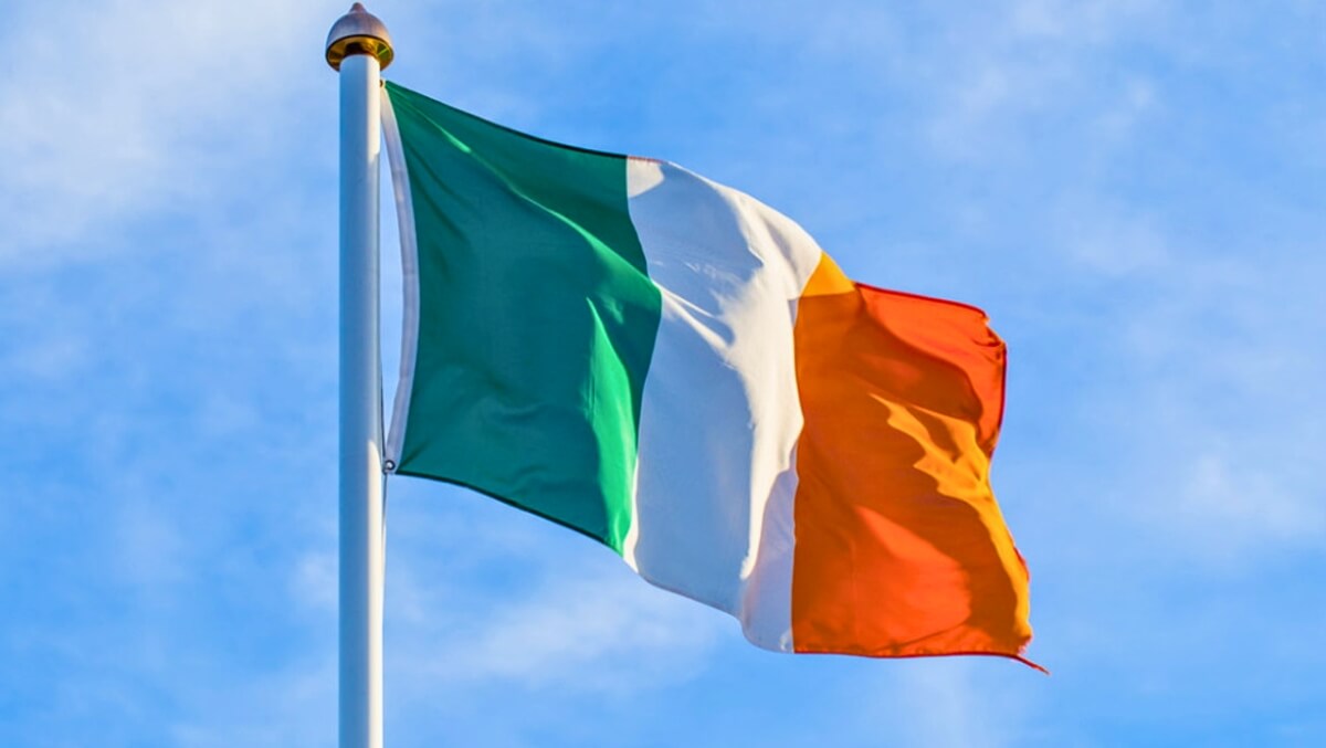 Irish flag with green, white, and orange stripes waving on a pole against a clear blue sky.