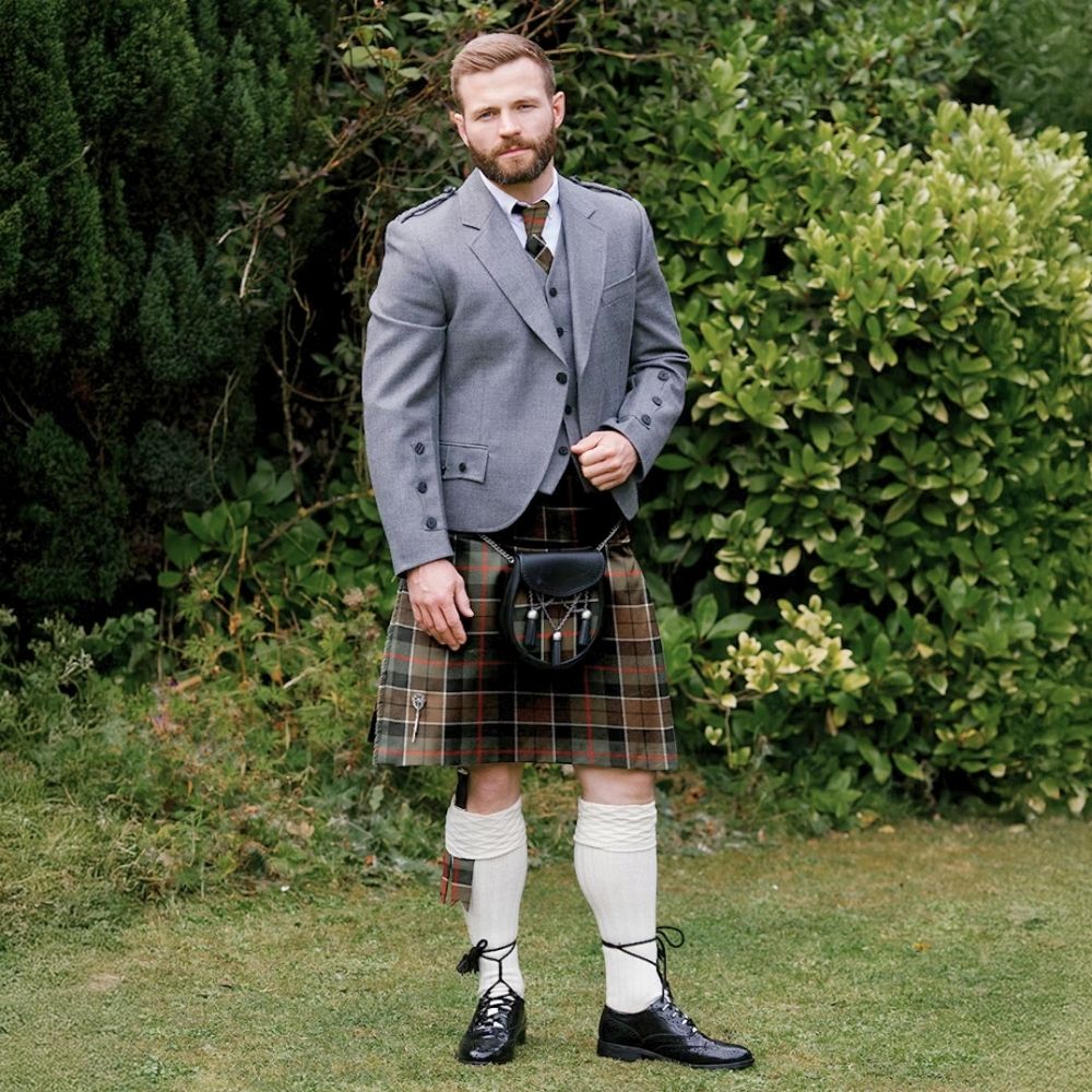 Man in Highland dress wearing a kilt, jacket, and front sporran, standing outdoors on a garden lawn.