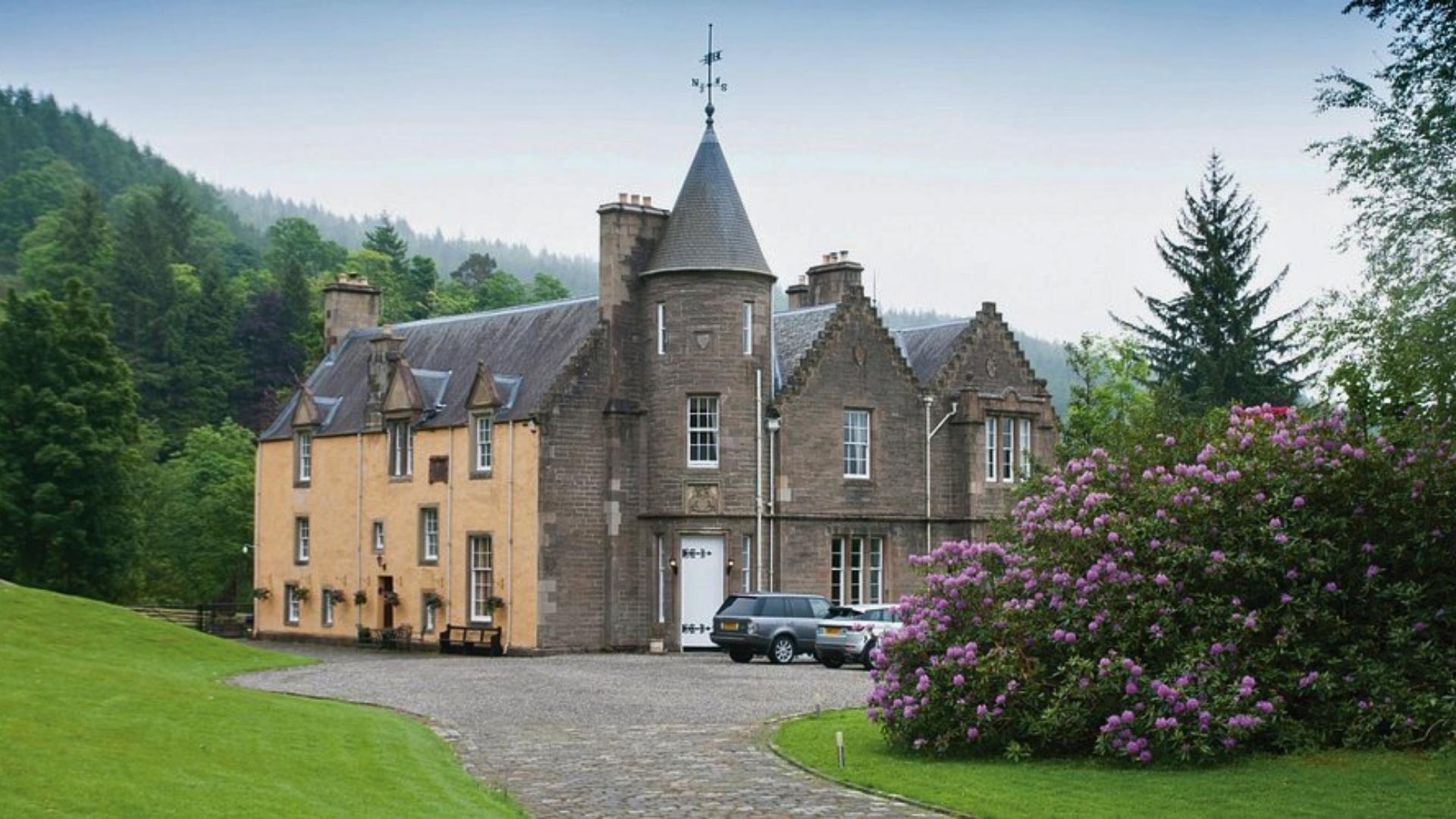 Large historic stone house with tower, set among trees, gardens, and parked cars in a lush Highland landscape.