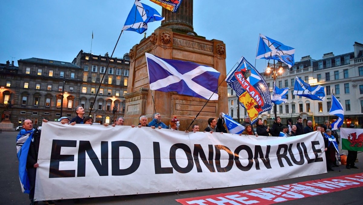 Crowd holding an “End London Rule” banner while waving Saltire flags during a pro-independence rally in a city square.