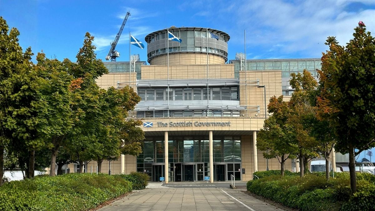 Front view of the Scottish Government building with Saltire flags flying on the rooftop and trees lining the walkway.