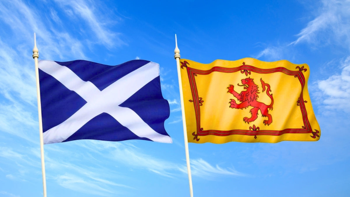 Saltire and Lion Rampant flags flying on tall poles against a bright blue sky with scattered clouds.