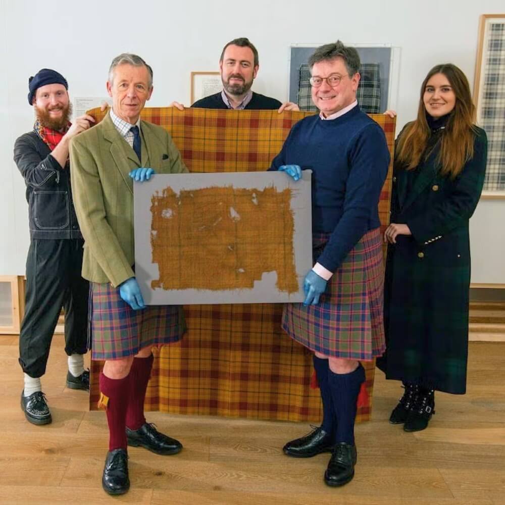 Group of people holding a fragile brown ancient textile with faint check lines, displayed in front of modern tartan fabrics.
