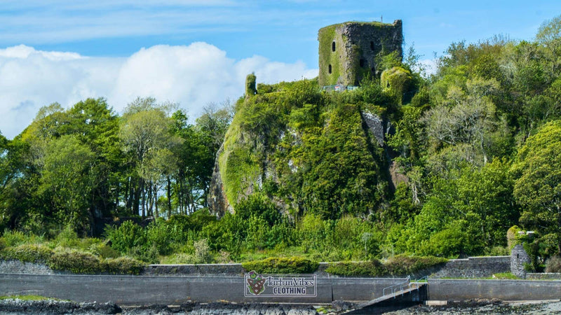 Close-up view of Dunollie Castle, a historic Scottish ruin covered in ivy, perched on a rocky green hill surrounded by dense trees and overlooking the Oban shoreline under a bright blue sky.