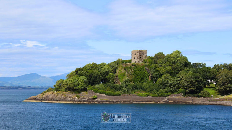 Dunollie Castle, a historic medieval ruin perched on a wooded hill overlooking Oban Bay in Scotland, surrounded by lush greenery and calm blue waters under a clear sky.