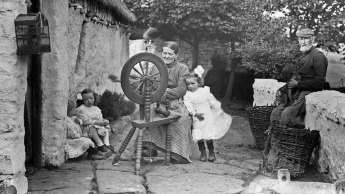 Vintage scene of a woman spinning wool on a wheel outdoors, with children and an older man nearby in a rural Irish setting.