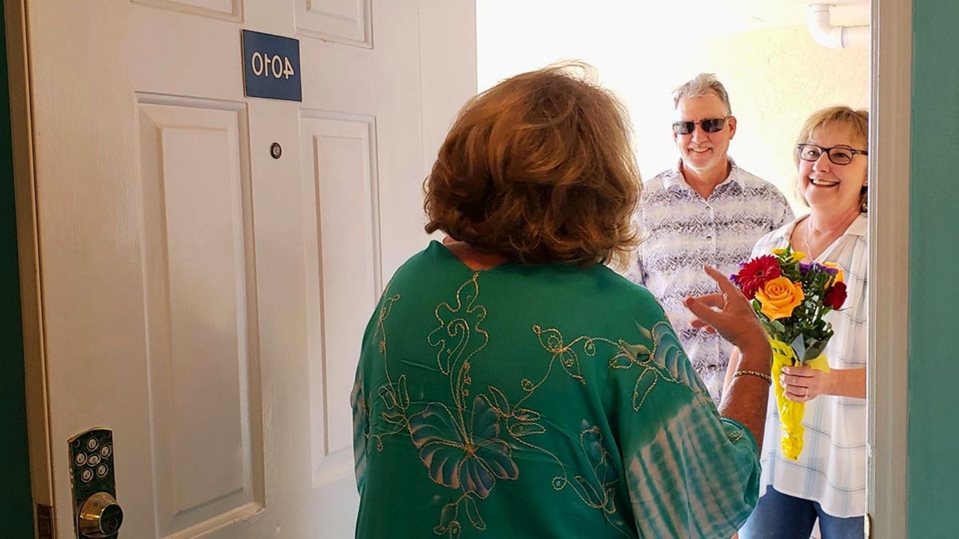 Woman opening her door to greet a smiling couple, with the woman holding a bouquet of flowers.