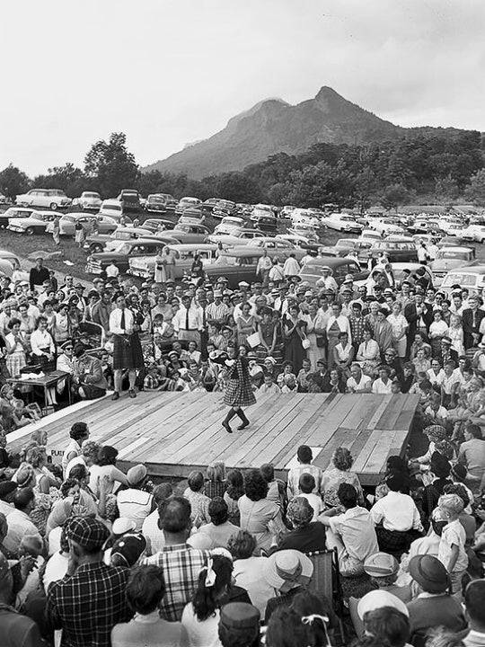 Girl in Scottish attire dancing on a wooden platform as a large crowd watches during the inaugural Grandfather Mountain Highland Games in 1956.