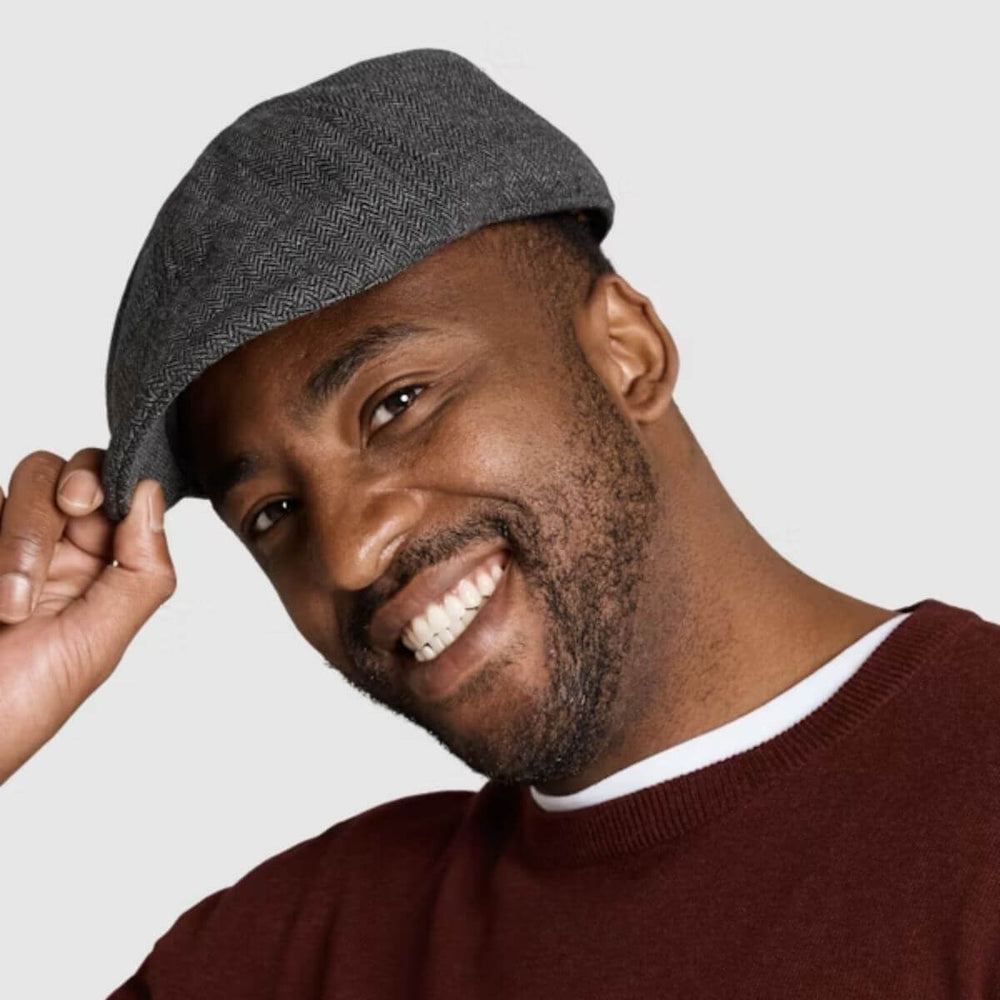 Smiling man wearing a grey herringbone flat cap against a light background