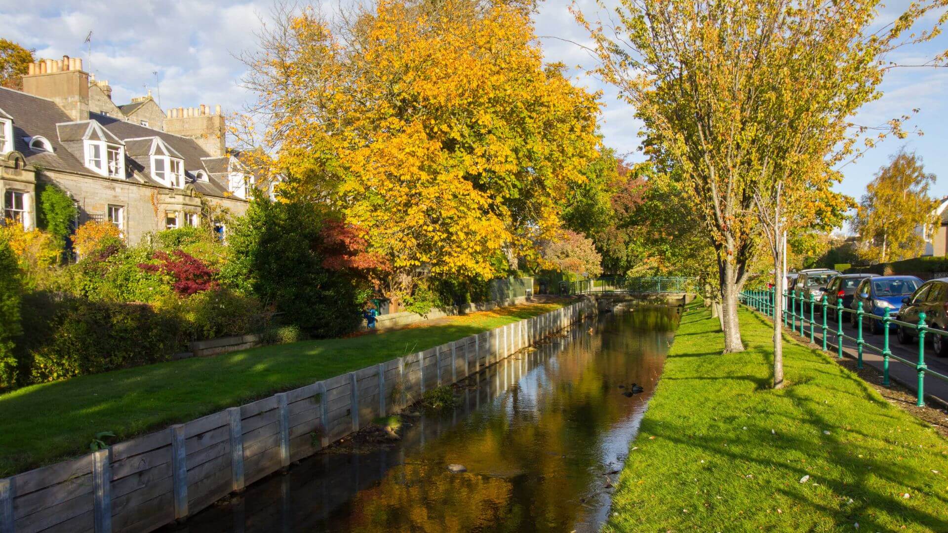 Scenic view of the Lade Braes Walkway in St Andrews, Scotland — featuring a tranquil stream, autumn-colored trees, and charming stone houses lining the peaceful path.