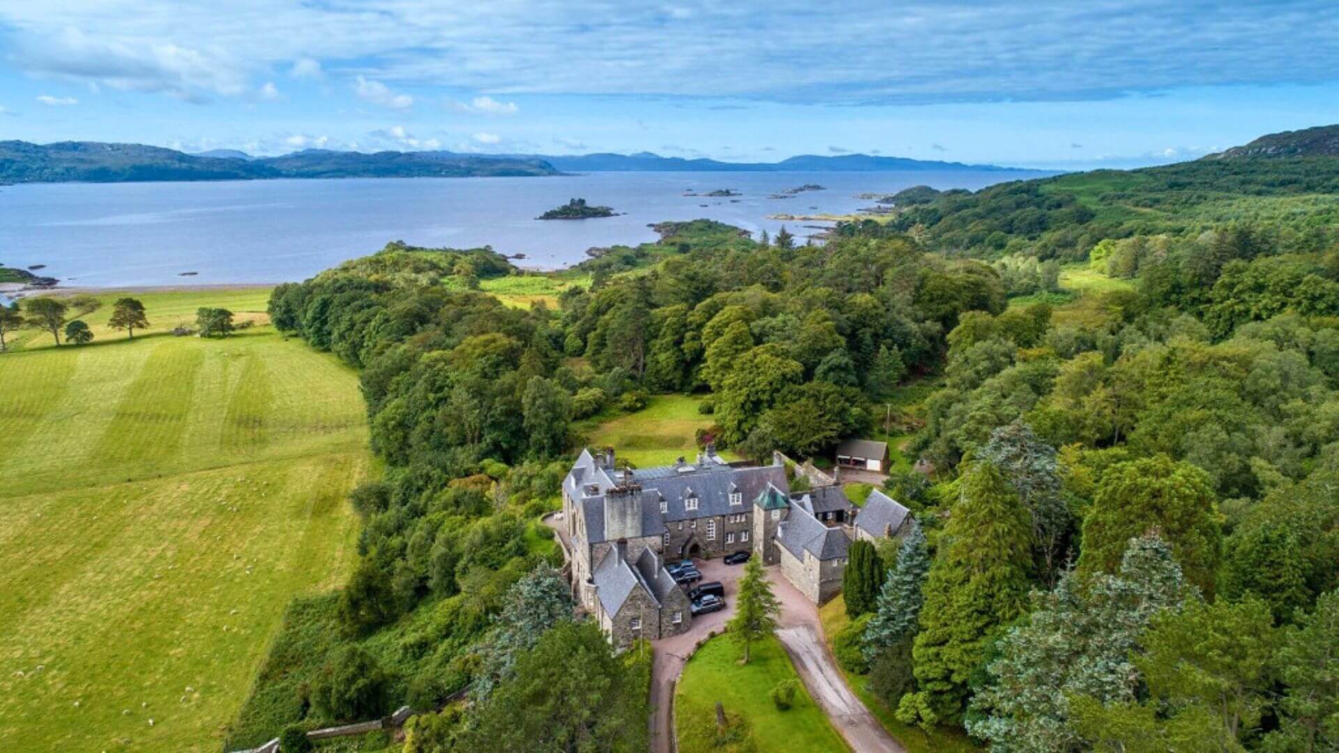 Aerial view of Lunga House, a historic Scottish mansion surrounded by lush greenery, fields, and overlooking a serene coastline with small islands and hills in the distance.