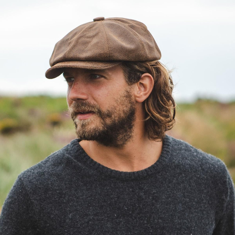 A man with a round face wearing a brown newsboy cap, looking to the side outdoors.