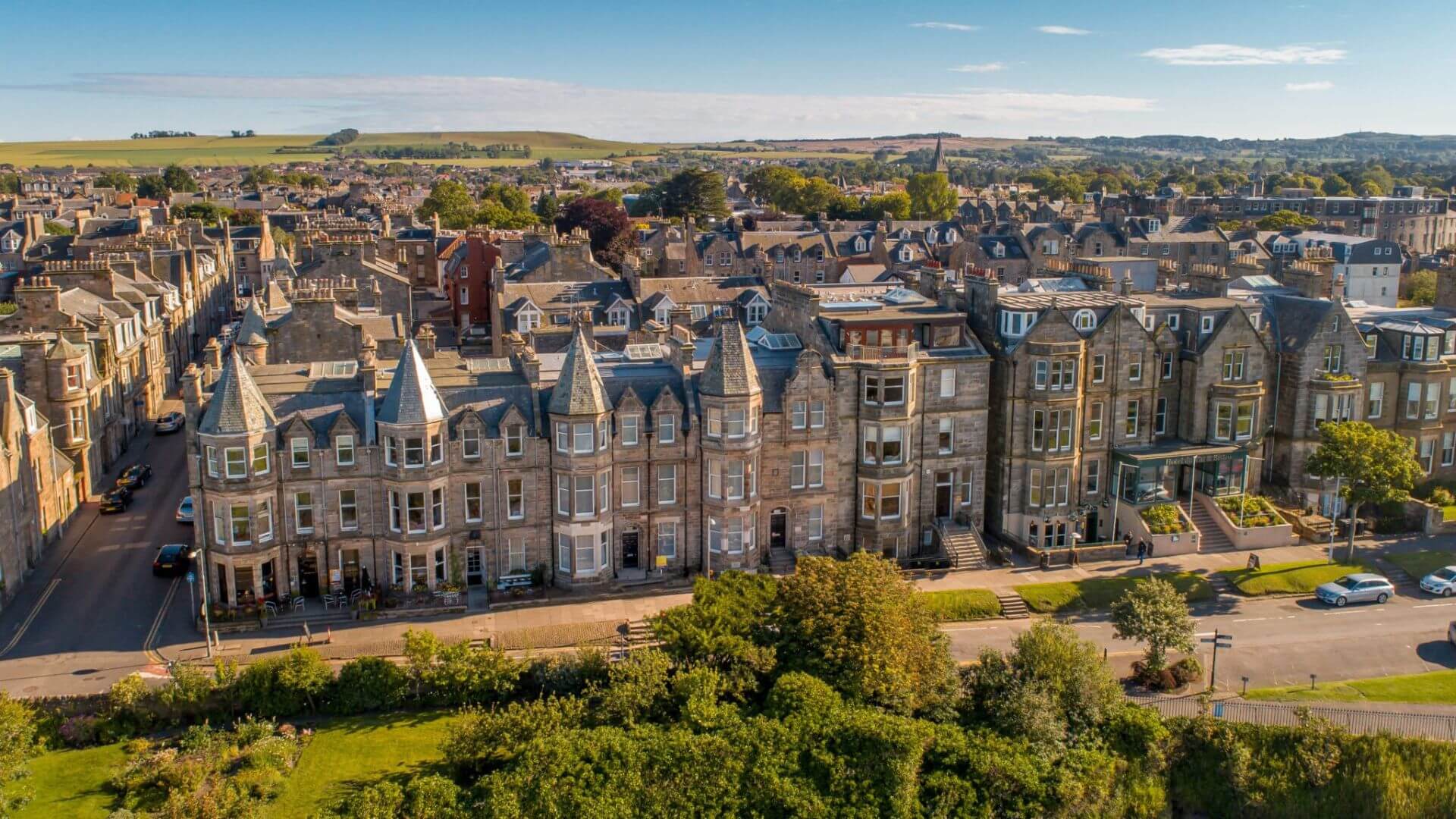 Aerial view of The Scores, St Andrews, Scotland — showcasing historic sandstone buildings, tree-lined streets, and the charming coastal townscape under a bright blue sky.